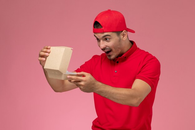 Front view of male courier in red uniform and cap holding little delivery package on pink wall