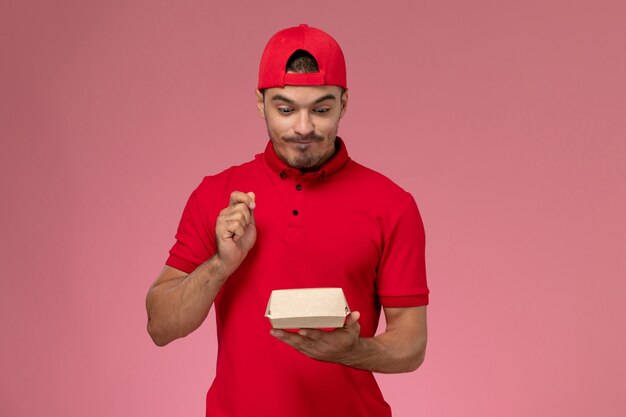 Front view of male courier in red uniform and cap holding little delivery package on pink wall