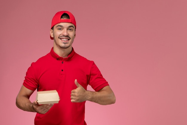 Front view of male courier in red uniform and cap holding little delivery package and smiling on pink wall