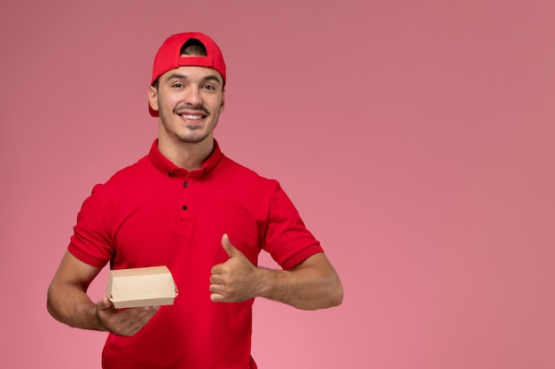 Вид спереди курьера-мужчины в красной форме и кепке, держащего небольшую посылку и улыбающегося на розовой стене