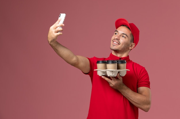 Front view male courier in red uniform and cape holding delivery coffee cups and taking photo on the pink wall 