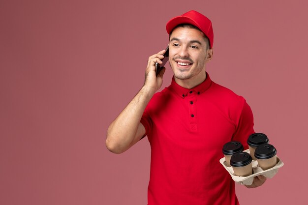 Front view male courier in red uniform and cape holding delivery coffee cups talking on the phone on pink wall 