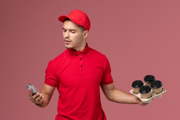 Курьер-мужчина, вид спереди в красной форме и накидке, держит кофейные чашки для доставки и использует свой телефон на розовой стене