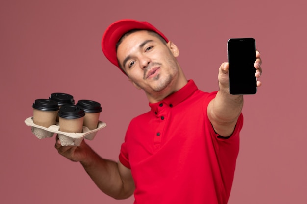 Corriere maschio di vista frontale in uniforme rossa e mantello che tiene le tazze di caffè di consegna con il telefono sulla parete rosa-chiaro