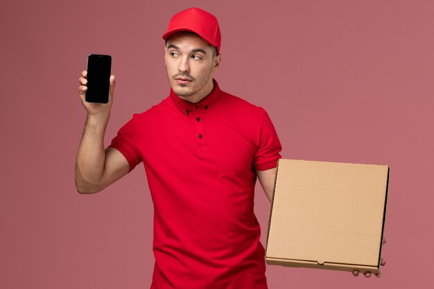 Front view male courier in red uniform and cape holding food box along with his phone on pink wall service job male delivery uniform