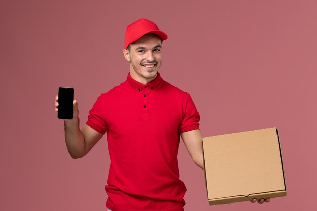 Front view male courier in red uniform and cape holding food box along with phone on pink wall service job male delivery uniform