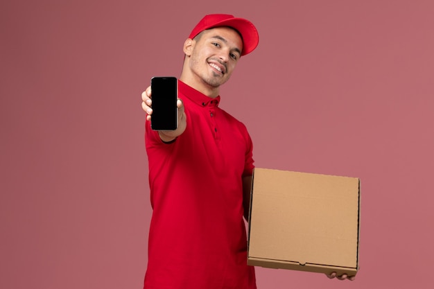 Foto gratuita corriere maschio di vista frontale in uniforme rossa e contenitore di cibo della tenuta del capo insieme al telefono che sorride sull'uniforme maschio di consegna di lavoro di servizio della parete rosa