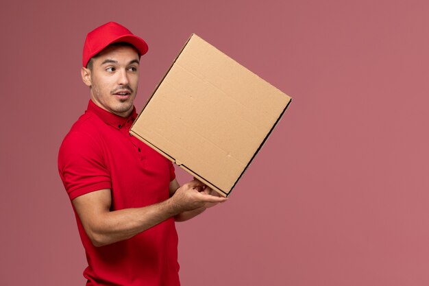 Front view male courier in red uniform and cape holding food box on light-pink wall worker job