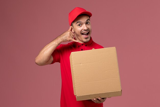 Front view male courier in red uniform and cape holding food box on light-pink wall worker