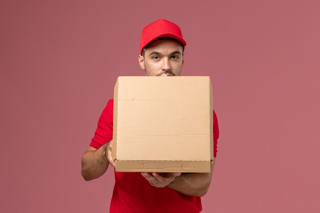 Front view male courier in red uniform and cape holding food box opening it on the light-pink desk worker job