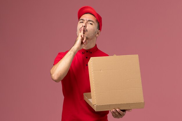 Front view male courier in red uniform and cape holding food box opening it on the pink wall worker job