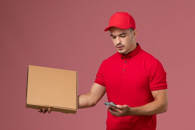 ピンクの壁のサービスの仕事の男性の配達の制服で彼の電話を使用してフードボックスを保持している赤い制服と岬の正面図男性宅配便