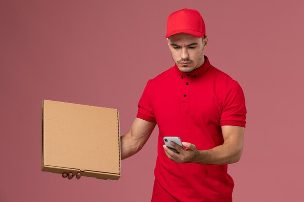 Курьер-мужчина, вид спереди в красной форме и плаще, держащий коробку с едой и использующий телефон на розовой стене, служба доставки мужской формы
