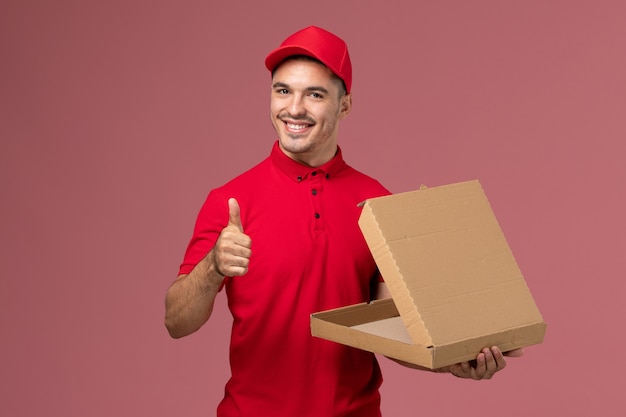 Курьер-мужчина в красной форме и накидке с улыбкой держит коробку для еды на розовом стенном рабочем, вид спереди