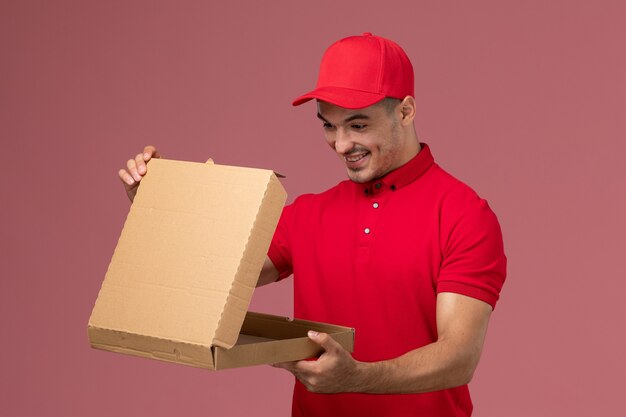 Front view male courier in red uniform and cape holding food delivery box opening it on pink wall service delivery male uniform worker