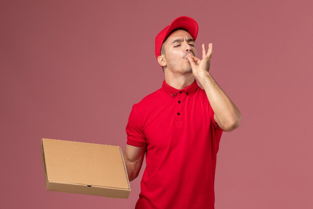 Курьер-мужчина в красной форме и плащ, держащий коробку для доставки еды на розовом полу, вид спереди