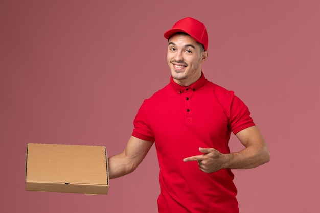 Курьер-мужчина, вид спереди в красной форме и плаще, держит коробку для доставки еды на розовой стене рабочего