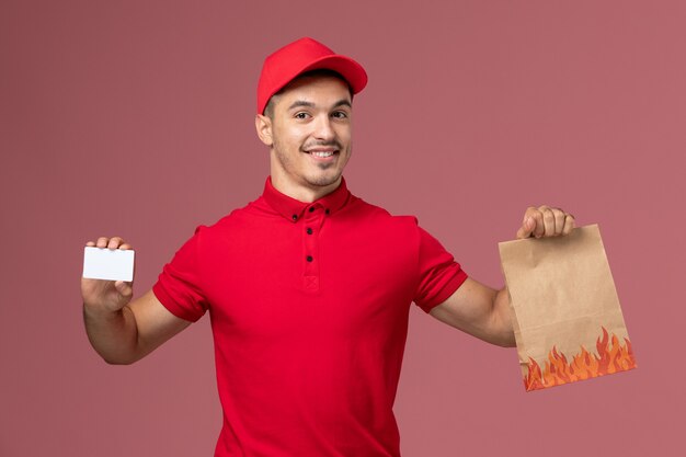 Front view male courier in red uniform and cape holding food package and card on the pink wall job