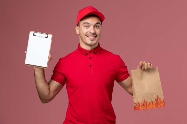 赤いユニフォームとピンクの壁のサービス配達労働者のユニフォームに食品パッケージとメモ帳を保持している岬の正面図男性宅配便