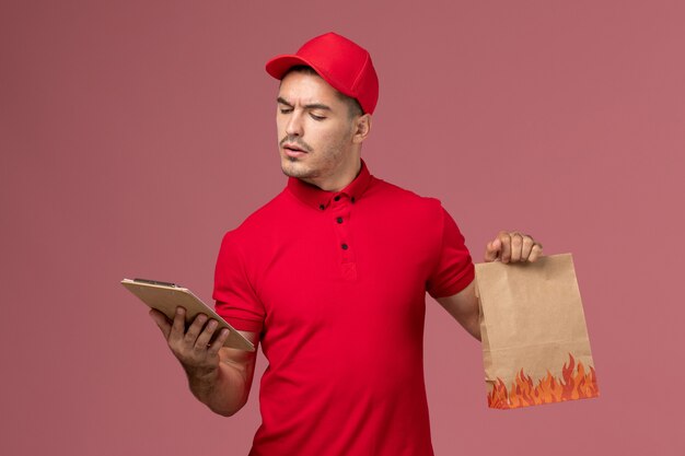 Курьер-мужчина в красной форме, вид спереди, держит пакет с едой и блокнот, читает его на розовой стене, рабочая форма работника службы доставки