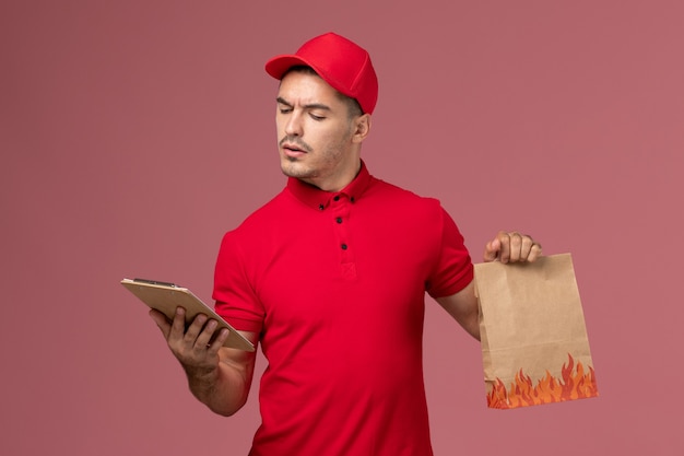 Front view male courier in red uniform and cape holding food package and notepad reading it on pink wall service delivery worker job uniform