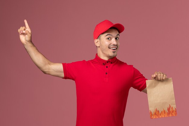Курьер-мужчина в красной форме и накидке, держащий пакет с едой на розовом стенном рабочем, вид спереди