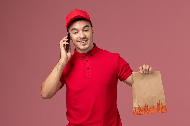 빨간색 유니폼과 케이프 음식 패키지를 들고 분홍색 벽에 전화로 이야기 전면보기 남성 택배