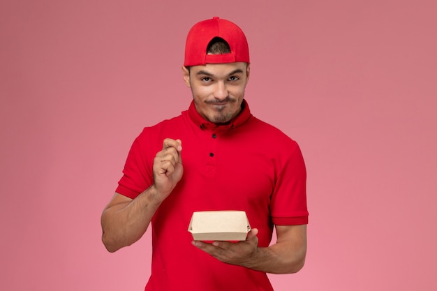 Front view male courier in red uniform and cape holding little delivery package on the pink background.