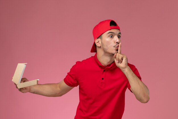 Front view male courier in red uniform and cape holding little delivery package on the pink background.