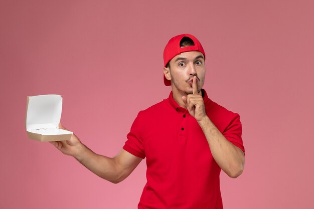 Front view male courier in red uniform and cape holding little delivery package on the pink desk.