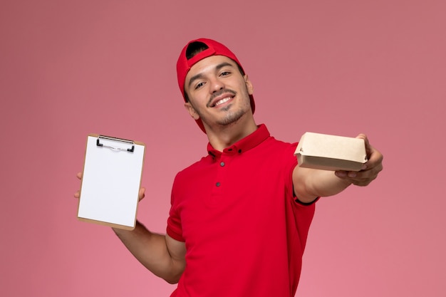 Курьер-мужчина вида спереди в красной форме и накидке, держащий маленький пакет доставки с блокнотом на розовом фоне.