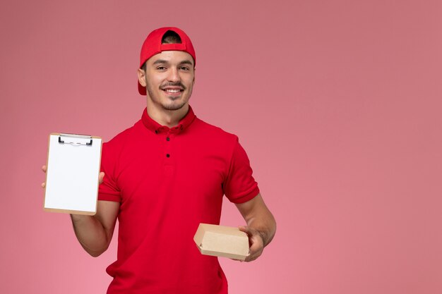 Front view male courier in red uniform and cape holding little delivery package with notepad on the pink background.