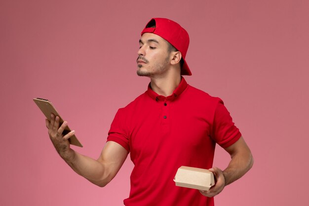 Front view male courier in red uniform and cape holding little delivery package with notepad on the pink background.