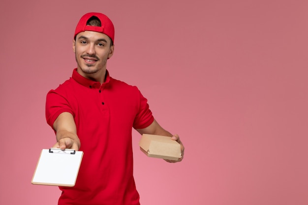 Front view male courier in red uniform and cape holding little delivery package with notepad smiling on the pink background.