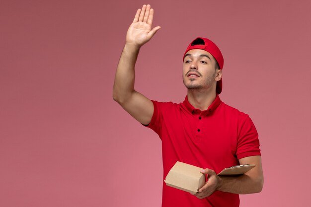 Front view male courier in red uniform and cape holding little delivery package with notepad waving on pink desk.