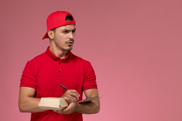 빨간색 유니폼과 케이프 분홍색 배경에 메모를 작성하는 메모장 작은 배달 패키지를 들고 전면보기 남성 택배.
