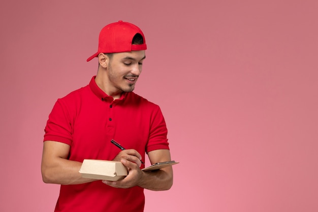 Front view male courier in red uniform and cape holding little delivery package with notepad writing note on pink background.