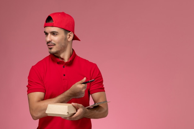 Front view male courier in red uniform and cape holding little delivery package with notepad writing note on pink background.