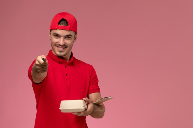 Corriere maschio di vista frontale in uniforme rossa e mantello che tiene piccolo pacchetto di consegna con la nota di scrittura del blocchetto per appunti che sorride su fondo rosa.