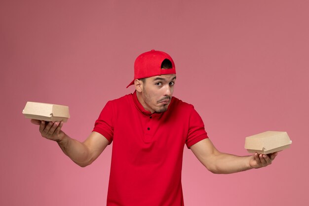 Front view male courier in red uniform and cape holding little delivery packages on the pink background.