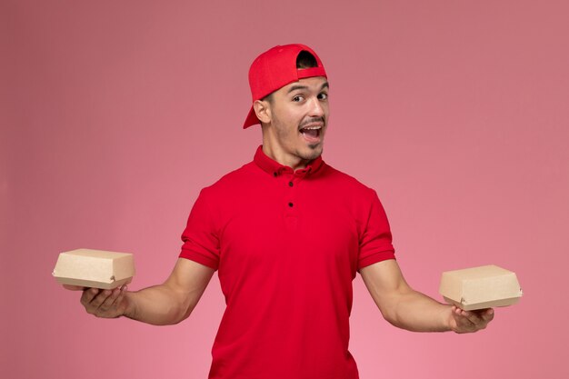 Front view male courier in red uniform and cape holding little delivery packages on the pink background.