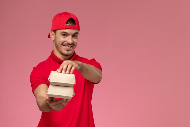 Corriere maschio di vista frontale in uniforme rossa e mantello che tiene piccoli pacchi di consegna sorridenti sui precedenti rosa.