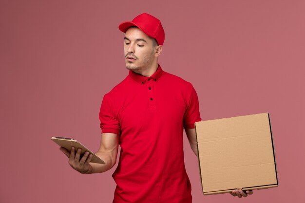 Front view male courier in red uniform and cape holding notepad food box on the pink wall service job male delivery uniform