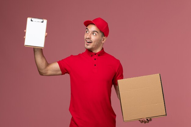 Front view male courier in red uniform and cape holding notepad and food box on pink wall 