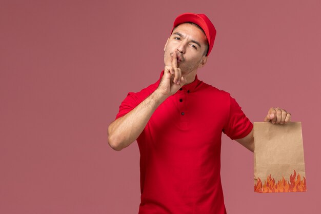 Курьер-мужчина в красной форме, вид спереди, держит бумажный пакет с едой и просит молчать о розовом стенном рабочем