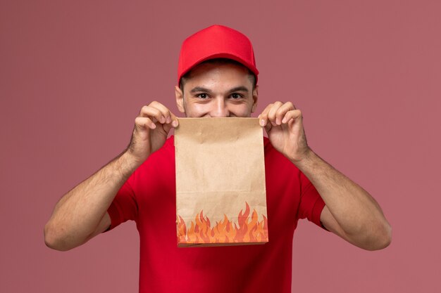 Front view male courier in red uniform and cape holding paper food package on pink wall service delivery male uniform worker