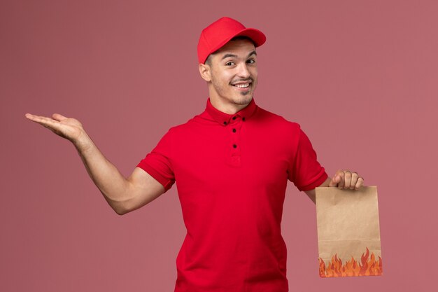 Front view male courier in red uniform and cape holding paper food package on pink wall 