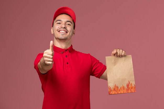 Front view male courier in red uniform and cape holding paper food package smiling on pink wall 