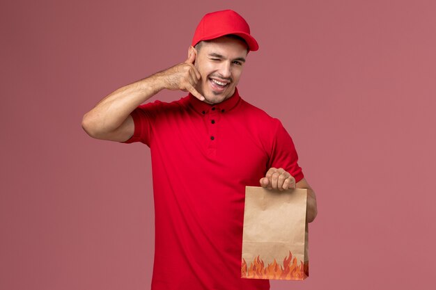 Front view male courier in red uniform and cape holding paper food package winking on the pink wall  worker