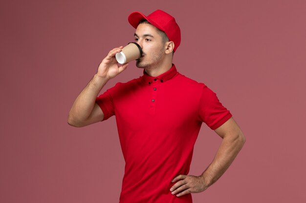Курьер-мужчина в красной форме пьет кофе на розовой стене